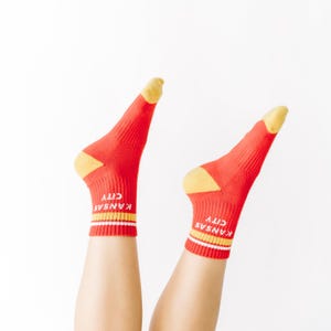 May include: A pair of red socks with yellow heels and toes. The socks feature the words "KANSAS CITY" in white. Yellow and white stripes are near the top of the socks.