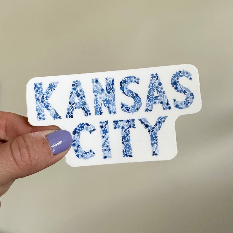 Peut inclure: Autocollant blanc avec les mots "Kansas City" en bleu. Les lettres sont compos&eacute;es de petits motifs floraux bleus.