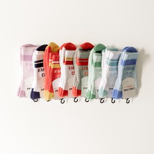 May include: A set of nine colorful socks with white stripes and the text "Kansas City" printed on them. The socks are hanging on a white background.
