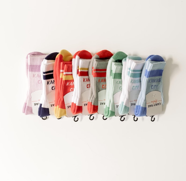 May include: A set of nine colorful socks with white stripes and the text "Kansas City" printed on them. The socks are hanging on a white background.