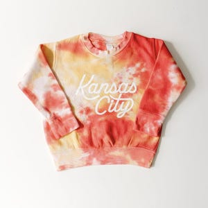 May include: A tie-dye sweatshirt in shades of red, orange, and yellow. The words "Kansas City" are printed in white script across the front. The sweatshirt has a crew neck and long sleeves.