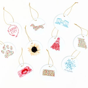 May include: Nine round ceramic ornaments with various designs, including a sunflower, a dachshund, a snowflake, a paw print, a red dress, a building, and the words "Kansas City".