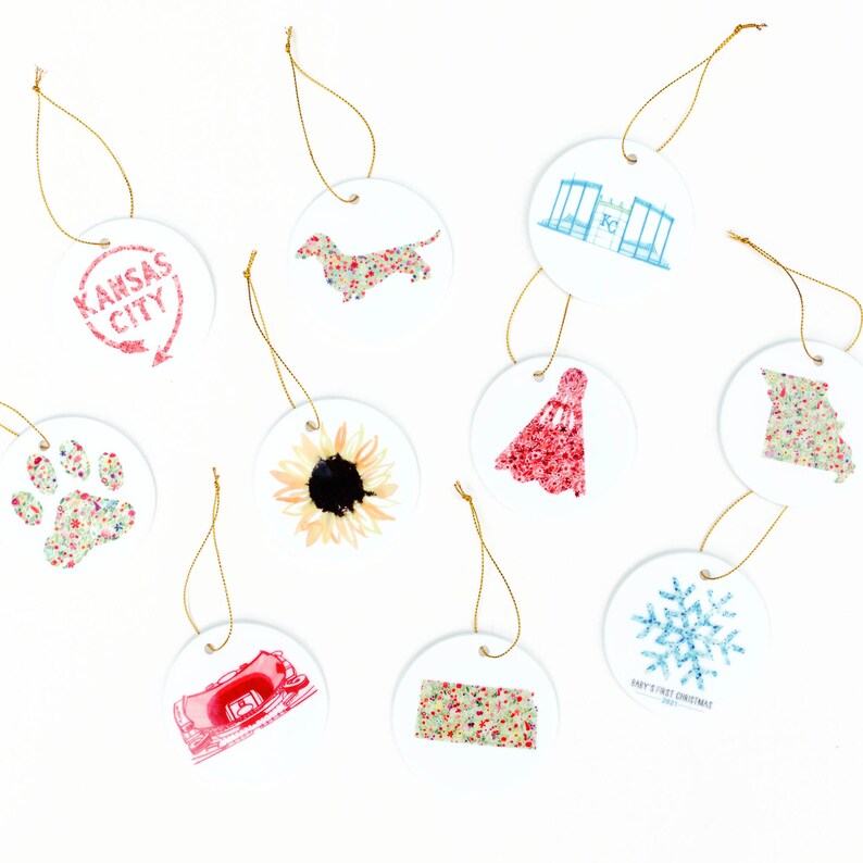 May include: Nine round ceramic ornaments with various designs, including a sunflower, a dachshund, a snowflake, a paw print, a red dress, a building, and the words "Kansas City".