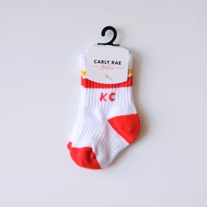 Peut inclure: Chaussettes de b&eacute;b&eacute; blanches avec des d&eacute;tails rouges sur le bout et le bord, avec les lettres "KC" en rouge. Le bord a des rayures jaunes. Les chaussettes sont attach&eacute;es &agrave; une &eacute;tiquette qui dit "CARLY RAE studio".