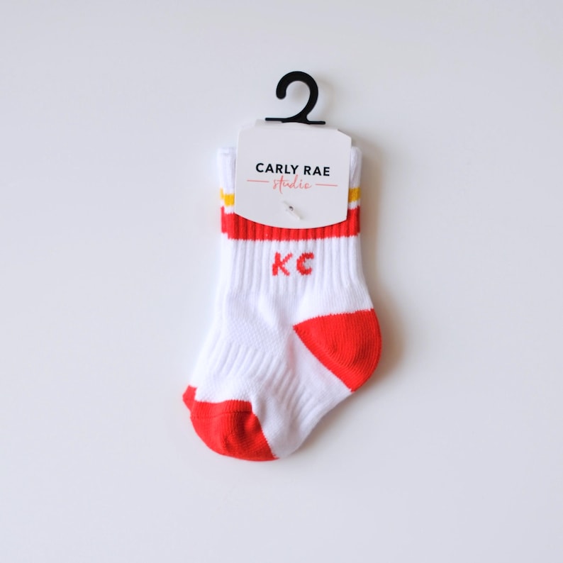 Peut inclure: Chaussettes de b&eacute;b&eacute; blanches avec des d&eacute;tails rouges sur le bout et le bord, avec les lettres "KC" en rouge. Le bord a des rayures jaunes. Les chaussettes sont attach&eacute;es &agrave; une &eacute;tiquette qui dit "CARLY RAE studio".