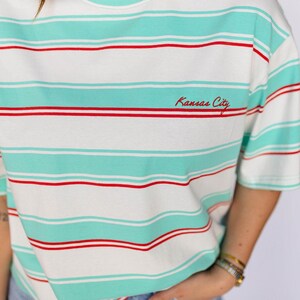 May include: White t-shirt with horizontal stripes in seafoam green and red. The text "Kansas City" is embroidered in red on the front. The shirt has short sleeves and a round neckline.