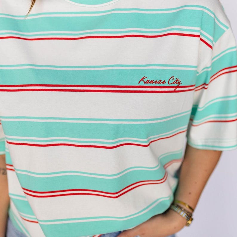 May include: White t-shirt with horizontal stripes in seafoam green and red. The text "Kansas City" is embroidered in red on the front. The shirt has short sleeves and a round neckline.