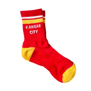 May include: Red crew socks with yellow heels and toes. The socks feature white and yellow stripes at the top and the words "KANSAS CITY" in white block letters.
