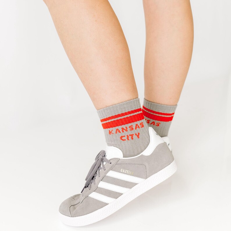 Op de afbeelding: Grijze enkelsokken met twee horizontale rode strepen aan de bovenkant en de tekst KANSAS CITY in rode letters. De sokken worden gedragen met grijze su&egrave;de sneakers met witte strepen en witte zolen, wat een klassiek sportief ontwerp toont.