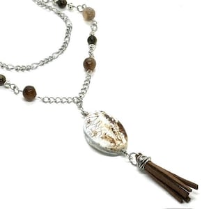 May include: A silver chain necklace with a large, oval pendant. The pendant is clear with a white and brown pattern. The necklace also features brown beads and a brown leather tassel. The necklace is a unique piece of jewelry.