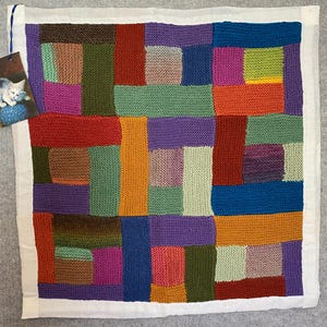 May include: A colourful knitted patchwork quilt with squares of various colours, including red, orange, yellow, green, blue, purple, brown, and pink.