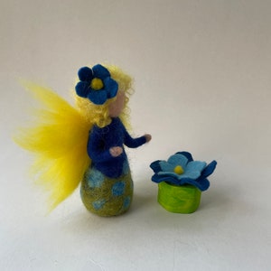 Op de afbeelding: Een vilten feepop met geel haar en blauwe vleugels, die een blauwe bloem vasthoudt. De fee draagt een blauwe jurk met groene en blauwe stippen. De bloem staat in een groene pot.