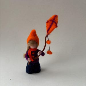 Könnte beinhalten: Eine nadelgefilzte Figur mit hellbraunem geflochtenem Haar, die einen leuchtend orangefarbenen spitzen Hut, eine violette Jacke mit orangefarbenen Knöpfen, einen roten Schal und einen dunkelblauen Rock trägt und einen orangefarbenen Drachen hält.