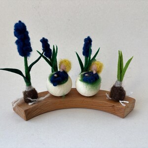 Op de afbeelding: Een houten display met vilten hyacintenbloemen in tinten blauw, groen en wit. De opstelling bevat verschillende hyacintenbloemen en -bollen, met kleine figuurtjes genesteld tussen de bloemen. De display staat op een gebogen houten voet.