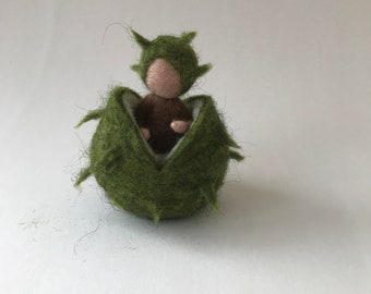 May include: A small felt doll with a green felt hat sits inside a green felt chestnut burr.