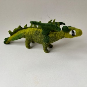 Könnte beinhalten: Handgefertigte Drachenfigur aus Filz in Grün- und Gelbtönen. Der Drache hat Flügel, einen langen Schwanz und kleine Beine. Der Drache ist ein skurriles Fantasy-Objekt.