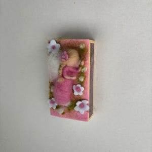 May include: A rectangular matchbox featuring a needle-felted scene on its top surface. A small figure with blonde hair in a braid wears a pink dress, lying amidst green fibres and white felt flowers with pink centres on a pink background.