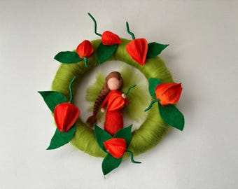 Op de afbeelding: Een handgemaakte vilten krans met een centrale figuur en bloemaccenten. De krans is heldergroen, versierd met feloranje bloemen en groene bladeren. In het midden staat een vilten pop in een rode jurk.