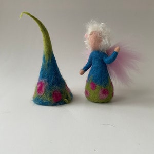 Op de afbeelding: Een kleine vilten feepop met wit haar, een blauwe jurk met roze bloemen en roze vleugels. De fee staat naast een blauwe en groene vilten hoed met roze bloemen.