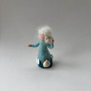 Könnte beinhalten: Eine nadelgefilzte Figur mit pfirsichfarbenem Gesicht und weißem Haar, die ein blaugrünes Kleid trägt, das in eine dunkelblaue Basis übergeht. Die Figur hält eine kleine Muschel am Ohr und drei weitere weiße Muscheln sind an der Basis befestigt.