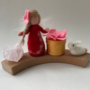May include: A felt doll with pink hair and a red dress stands on a wooden base with a pink crystal, a small white rabbit, and a pink flower in a gold box.