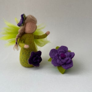 May include: A green felt fairy doll with a purple flower crown and a purple flower in her hand. The fairy is standing on a white background with a purple felt flower.