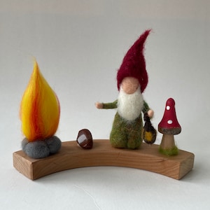 May include: A felt gnome with a red hat and green tunic stands next to a felt flame and a red and white mushroom. The gnome is holding a small lantern. All three figures are on a wooden base.