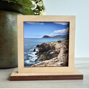 May include: A small, square wooden picture frame with a natural finish. The frame holds a scenic photograph of a coastal landscape with blue water, rocks, and a clear sky. The base of the frame is a darker wood tone. Text at the bottom reads "Natural Finish, with Solid Corners."