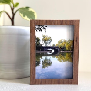May include: A wooden picture frame displaying a scenic photograph of a bridge over a calm body of water. The frame is a warm brown color and sits on a white surface. A potted plant with green and white leaves is in the background.