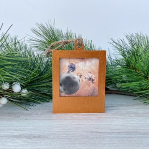 May include: A square wooden ornament with a gold finish. The ornament features a picture of a Christmas tree with ornaments. The ornament has a twine hanger.