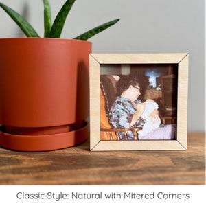 May include: A square wooden picture frame with mitered corners containing a photograph of a person sitting in a wooden rocking chair while holding a baby. The frame is light-colored wood and features the text Classic Style: Natural with Mitered Corners.
