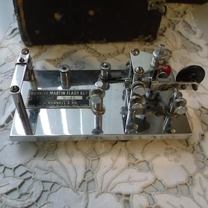 May include: A vintage Burnett Martin Flash Key telegraph transmitter featuring a polished chrome rectangular base, intricate mechanical components, and a black identification plate with white text reading Burnett Martin Flash Key.