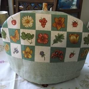 May include: A tea cozy with a green and white checkered base and a top featuring a patchwork design of flowers, fruits, and insects. The cozy has a loop handle and is set on a white tablecloth.
