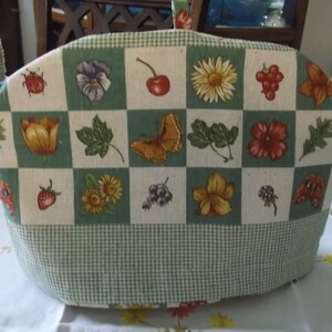 May include: A tea cozy with a green and white checkered pattern at the base and a top section featuring a grid of botanical illustrations. The illustrations include flowers, fruits, and insects.