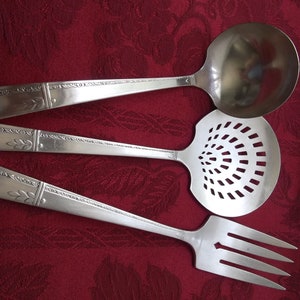 May include: Silver serving utensils set including a ladle, slotted spoon, and serving fork. Each piece has a decorative leaf design on the handle. The utensils are arranged on a red tablecloth.