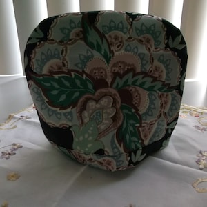 May include: A black fabric cover with a floral pattern in shades of green, blue, and pink. The cover has a rounded shape and is designed to fit over a small appliance.