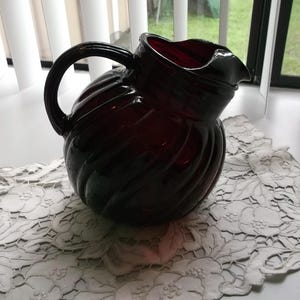 May include: A deep ruby red glass pitcher featuring a rounded, bulbous body with prominent vertical ribbed detailing. The vessel includes a curved handle on the side and a distinct pouring spout at the top rim, crafted from translucent colored glass.