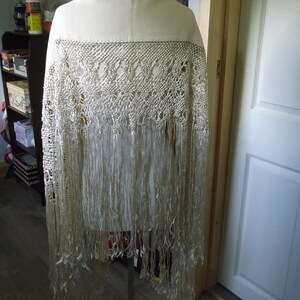 May include: A cream-colored shawl with a detailed, openwork design across the top and long fringe. The shawl is displayed on a mannequin, showcasing its texture and drape. The background includes a wooden door and a shelf.