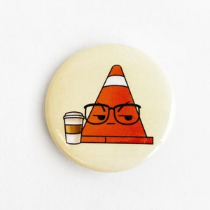 Peut inclure: Un bouton rond de couleur crème représentant un cône de signalisation orange portant des lunettes et l'air renfrogné. Une tasse de café est à côté du cône. Le bouton a un style de dessin animé et est probablement une épingle ou un badge.