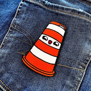 May include: A colourful patch featuring a cartoon traffic cone design, with orange and white stripes and a smiling face. The patch is sewn onto the back pocket of blue denim jeans, a playful accessory.