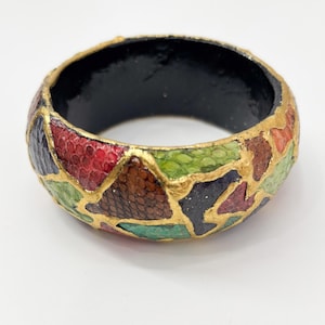 May include: A chunky, colorful bangle bracelet. The bracelet features a mosaic pattern of red, green, brown, and black segments, outlined with gold. The interior is black.