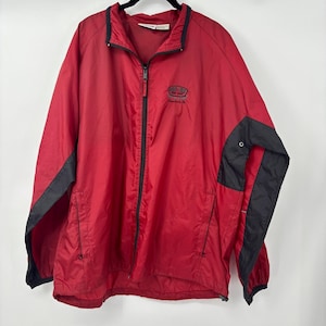 May include: A red zip-up jacket with black accents on the sleeves and collar. The jacket has a small embroidered logo on the chest and a full-length zipper. The jacket is made of a lightweight material.
