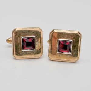 May include: A pair of square gold-tone cufflinks, each featuring a central square-cut deep red gemstone set within a silver-tone border. The metal surface has a subtle hammered texture, and the design includes a standard toggle backing mechanism.