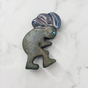 May include: A Kokopelli figure in a metallic finish, with a turquoise-colored eye. The figure is in a dancing pose, with a headpiece. The color palette includes shades of green, bronze, and blue.