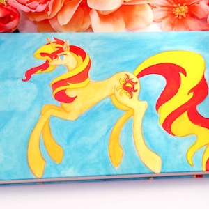 Könnte beinhalten: Ein hellblaues Notizbuch mit einem gelben Pony mit roter und gelber Mähne und Schweif. Das Pony hat ein Sonnenemblem an der Flanke. Das Notizbuch ist von rosa und orangefarbenen Blumen umgeben.