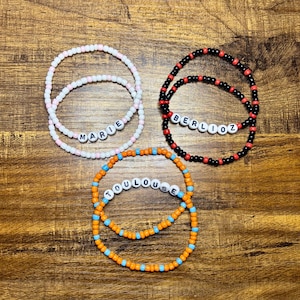 May include: Three beaded bracelets with names. One bracelet is white and pink with the name "MARIE". Another is black and red with the name "BERLIOZ". The third is orange and blue with the name "TOULOUSE".