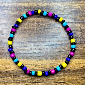 May include: A beaded bracelet featuring a colorful pattern of black, yellow, blue, cyan, and magenta beads. The bracelet is strung on a clear elastic cord and is displayed on a wooden surface.