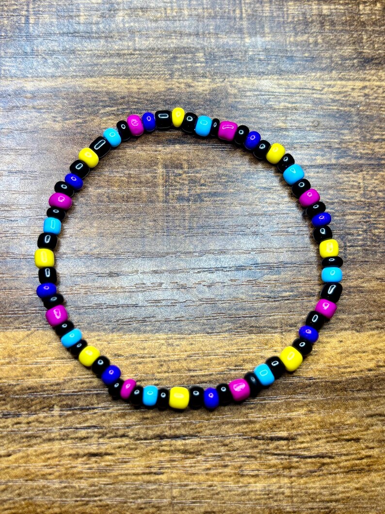 May include: A beaded bracelet featuring a colorful pattern of black, yellow, blue, cyan, and magenta beads. The bracelet is strung on a clear elastic cord and is displayed on a wooden surface.