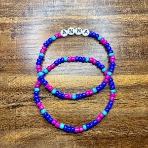 May include: Two beaded bracelets. The top bracelet has the name "ANNA" spelled out in white letter beads. The bracelets are made of blue, pink, and turquoise beads. The bracelets are on a wooden surface.