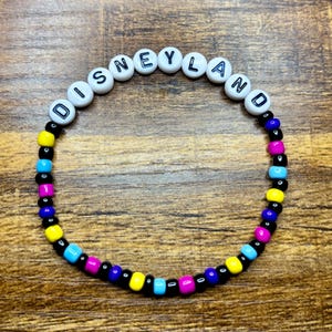 May include: A beaded bracelet with the word "DISNEYLAND" spelled out in white letter beads. The bracelet features a colorful pattern of black, yellow, blue, and pink beads, creating a vibrant accessory.